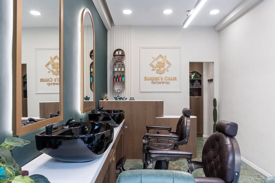 Το εσωτερικό του Barber's Chair: το χρυσό σήμα στον τοίχο, δύο μαύροι νιπτήρες, ξύλινοι καθρέφτες σε καμάρα με κρυφό φωτισμό και δερμάτινες καρέκλες σε πράσινο δάπεδο