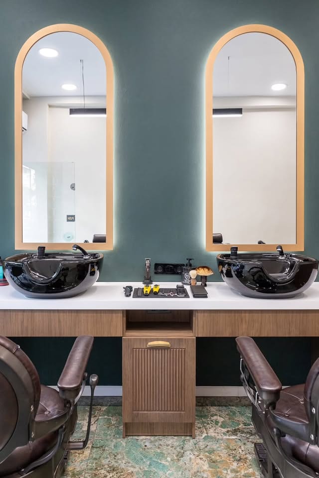 Οι δύο καθρέφτες σε καμάρα του Barber's Chair, συμμετρικοί, με κρυφό φωτισμό, οι μαύροι νιπτήρες και τα εργαλεία στον λευκό πάγκο